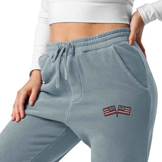 A pair of blue pigment-dyed sweatpants with an embroidered American flag on the left leg, featuring a drawstring waist and front pockets.